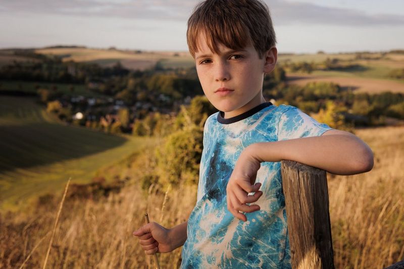 A child in a field resting his arm on a wooden post. A diffusion panel has been used to soften the sunlight. Taken on a Canon EOS R6 with a Canon RF 50mm F1.8 STM lens.