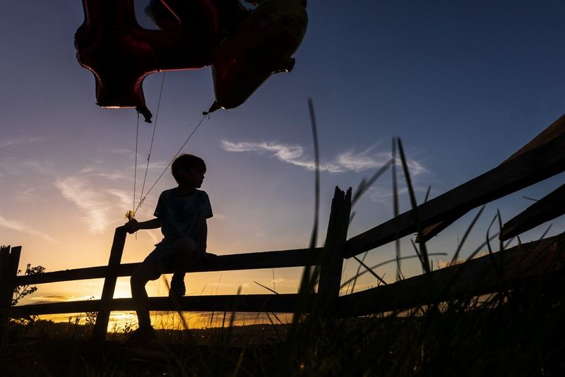 A child sitting on a fence holding a bunch of balloons silhouetted against the setting sun. Taken on a Canon EOS R6 with a Canon RF 24-105mm F4-7.1 IS STM lens.