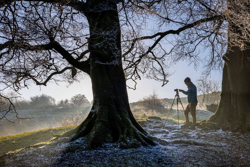 A man stands in a forest on a wintry day, with his Canon camera set up on a tripod. 