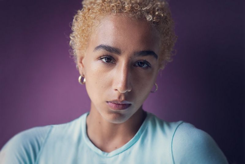 A portrait, lit from the side and taken on a Canon EOS R6 with a Canon RF 50mm F1.8 STM lens, of a person with curly blonde hair looking directly at the camera. 