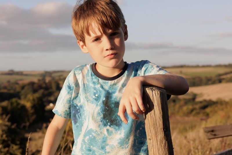 A child in a field resting his arm on a wooden post. The direct sunlight is casting harsh shadows on his face. Taken on a Canon EOS R6 with a Canon RF 50mm F1.8 STM lens.