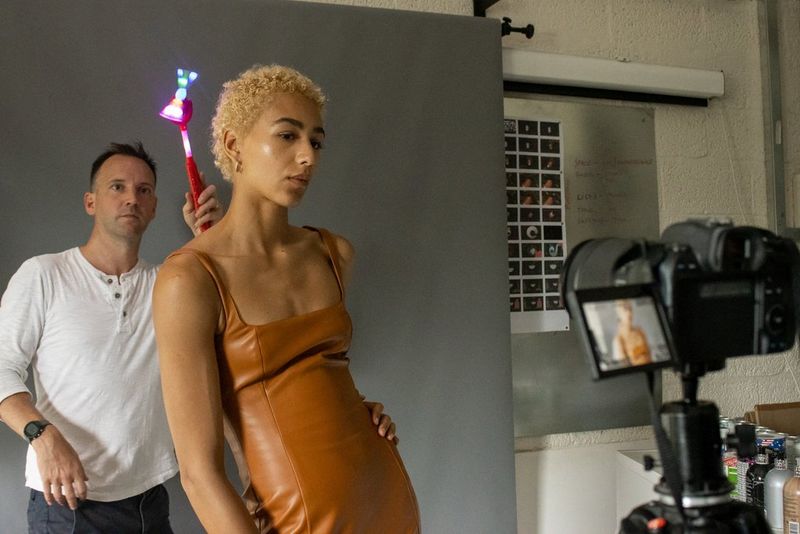 A woman poses in front of a Canon camera while a man holds a light behind her head. 