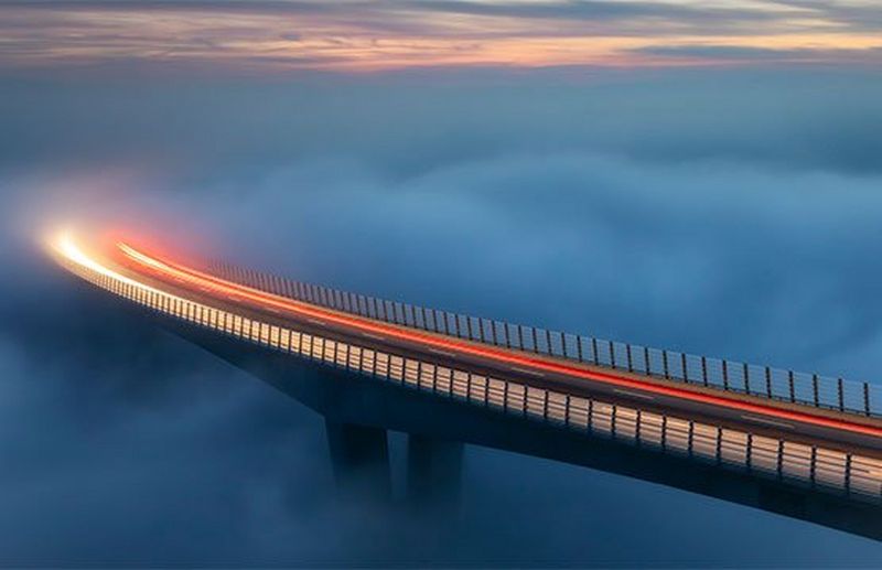 Car light trails in evening light across a curved bridge. Thick, cloud-like fog obscures the bridge supports and the ground below.