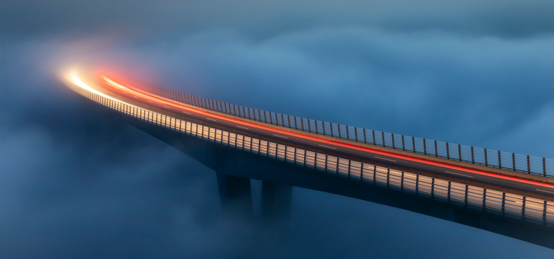 Car light trails in evening light across a curved bridge. Thick, cloud-like fog obscures the bridge supports and the ground below.