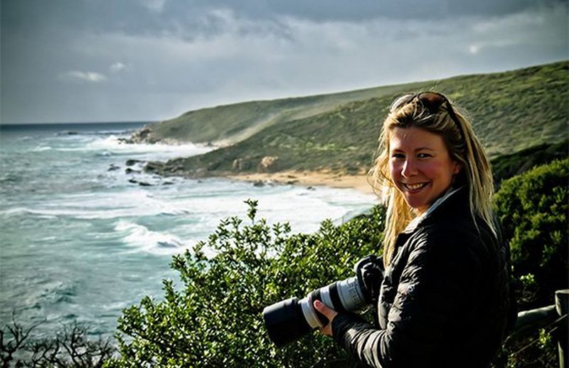 Photographer Camilla Rutherford smiles for the camera, waves lapping the shoreline behind her.