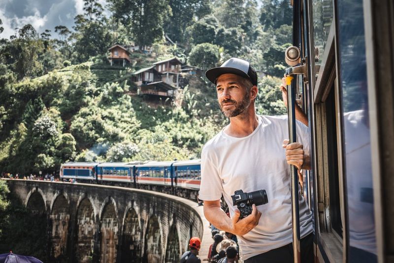 Fotograf Martin Bissig se nagiba iz vlaka, ki se je ustavil in pobira potnike na viaduktu na Šrilanki. Drži Canon EOS R10, za njim pa je grič z majhnimi lesenimi hišami. Fotografirala Monika Bissig s fotoaparatom Canon EOS R7. 