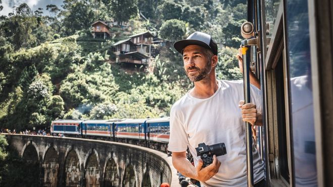 Fotograf Martin Bissig wychyla się z pociągu, który zatrzymał się na wiadukcie na Sri Lance, aby zabrać pasażerów. W dłoni trzyma aparat Canon EOS R10. Za nim widać wzgórze, na którym zbudowano małe drewniane domy. Zdjęcie zrobione przez Monikę Bissig aparatem Canon EOS R7. 