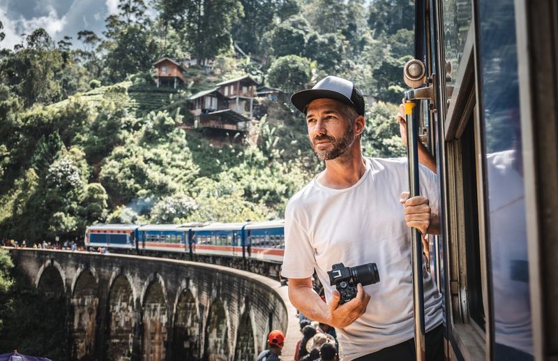 Photographer Martin Bissig leans out of a train that has stopped to pick up passengers on a viaduct in Sri Lanka. In his hand is a Canon EOS R10 and behind him a hill with small wooden houses built into it. Photographed by Monika Bissig on a Canon EOS R7. 