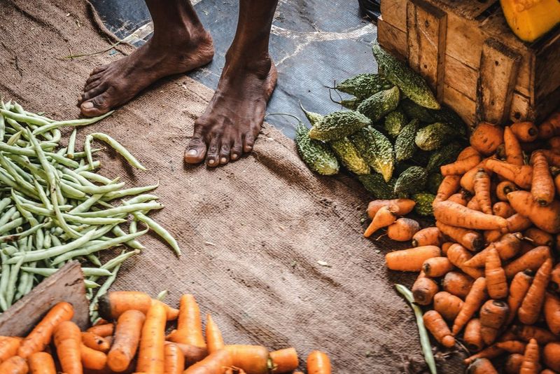 A close-up of bare feet next to small piles of vegetables including carrots and runner beans at a Sri Lankan market stall. Taken by photographer Martin Bissig on a Canon EOS R10.