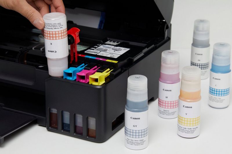 A hand pouring a bottle of red ink into a PIXMA G series printer with other bottles beside it.