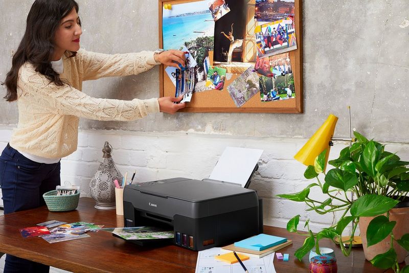 A woman pinning photo prints to a noticeboard. A Canon PIXMA G3520 printer is on the desk below.