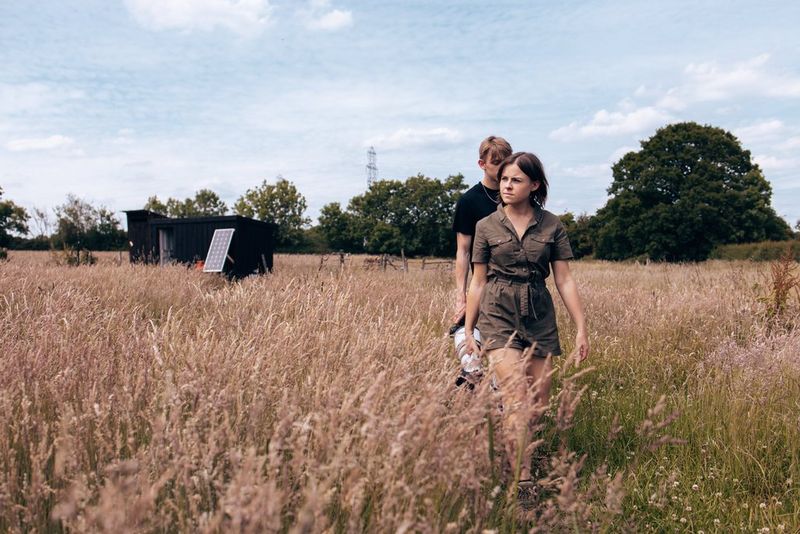 A young woman and man walk in single file through a field of long grass. In the background, a small bird hide can be seen.
