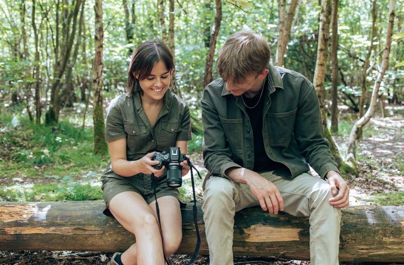 A young woman and man sitting on a wooden log in a forest setting smile happily as they study a camera touchscreen.