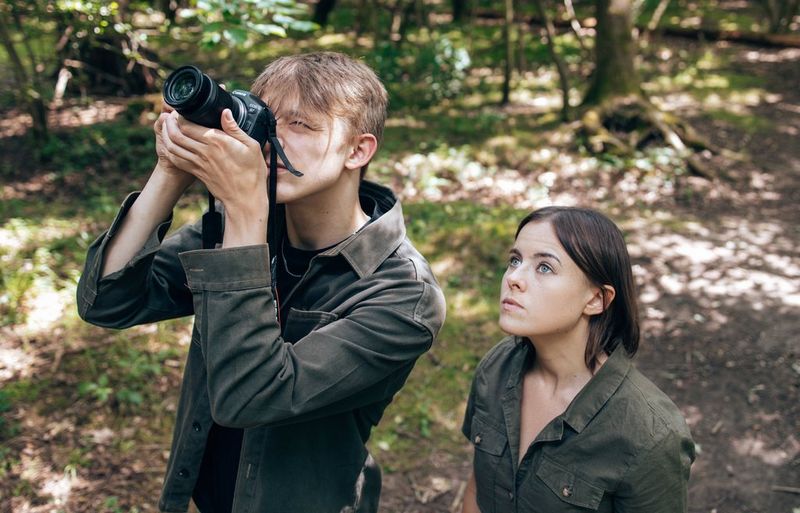 In a forest setting, a man takes a picture of something above him with a Canon camera and lens, as a woman watches, also looking up.