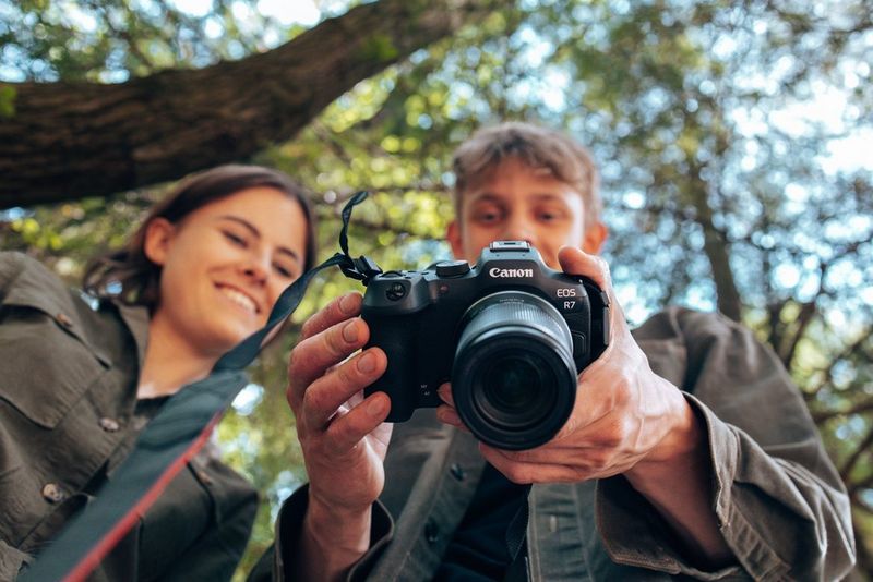A Canon camera and lens taken from below, with two smiling young people slightly blurred in the background looking at the touchscreen.