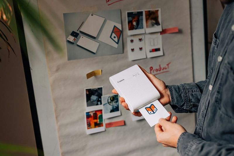A person holding a Canon SELPHY Square QX10 as it prints out a logo for a project. 