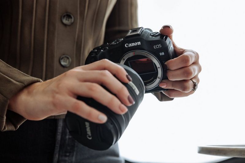 A pair of hands attaches a Canon lens to a Canon EOS R8 camera.