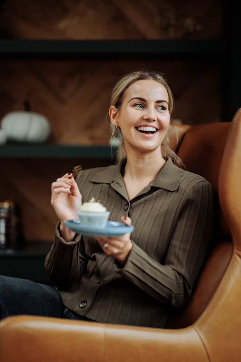 A woman sits on a sofa, smiling and holding a fork up to a cupcake on a plate in front of her.