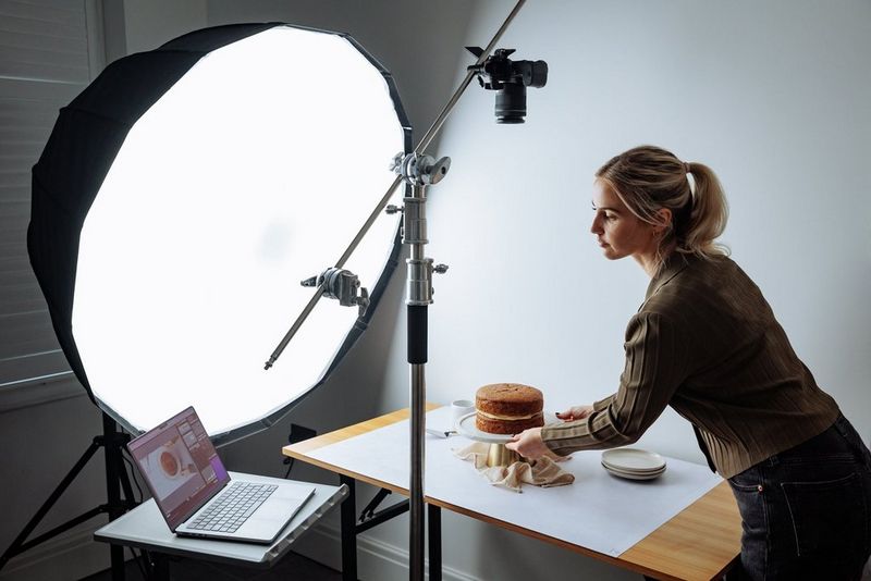 A woman arranges a cake in front of a softbox light, a laptop on a table nearby and a Canon EOS R8 camera positioned on a tripod to film from above.