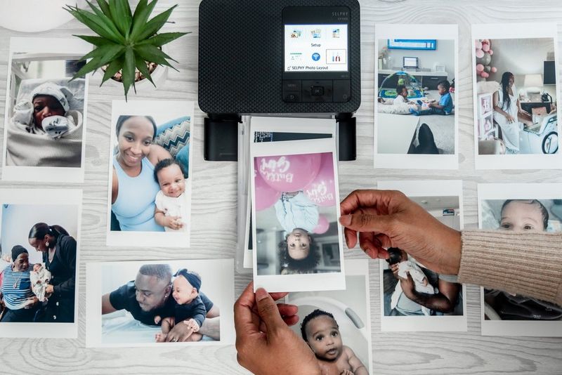 A pair of hands holds a printed baby photo above a Canon SELPHY CP1500, with further printed photos surrounding it.