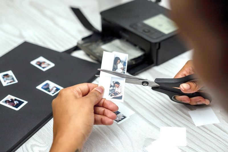 A person uses a pair of scissors to cut out printed images, preparing to stick them in their scrapbook.