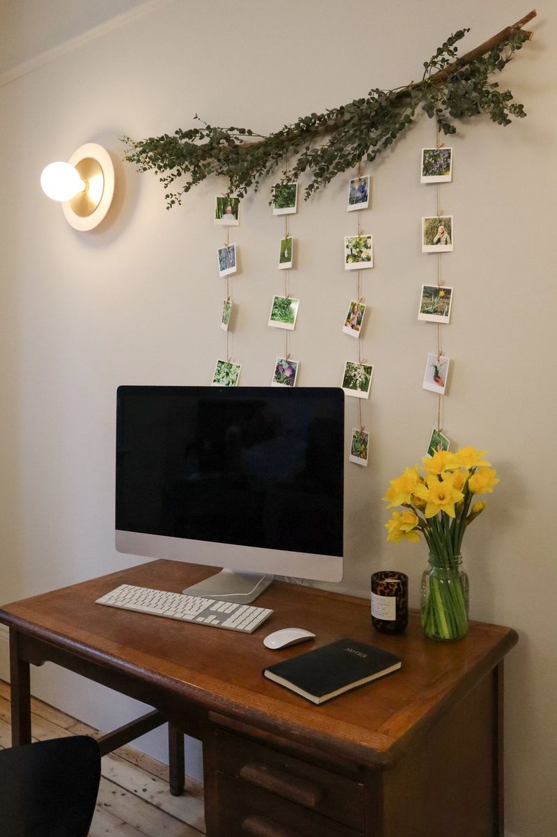A branch with small printed photographs hanging from it is positioned above a desk containing a computer, notebook, candle and vase of daffodils.