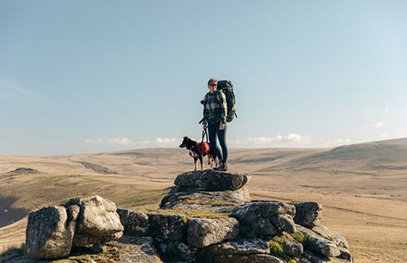 A person wearing hiking gear and a large rucksack stands on top of a rocky outcrop with their dog. In the background, acres of remote moorland can be seen.