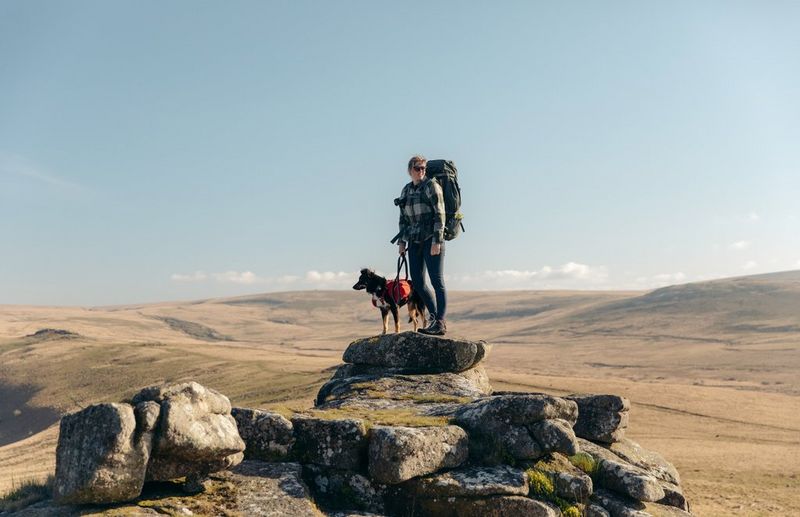 A person wearing hiking gear and a large rucksack stands on top of a rocky outcrop with their dog. In the background, acres of remote moorland can be seen.