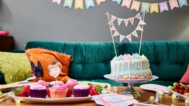 A large cake and smaller cupcakes are adorned with papercraft decorations and arranged on a coffee table next to photo props, with a papercraft banner hanging in the background.