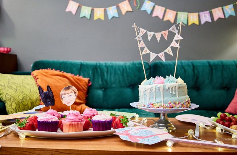 A large cake and smaller cupcakes are adorned with papercraft decorations and arranged on a coffee table next to photo props, with a papercraft banner hanging in the background. 