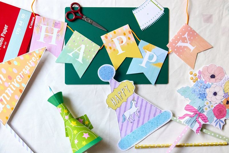 A range of birthday papercraft decorations, including a banner and numerous photo props, are spread across a table next to a pair of scissors, a cutting board and a pack of Canon paper.