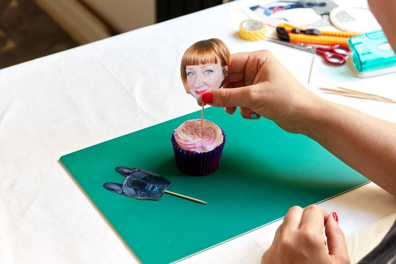 A hand places a food pick with an image of Pippa's head into the top of a cupcake.