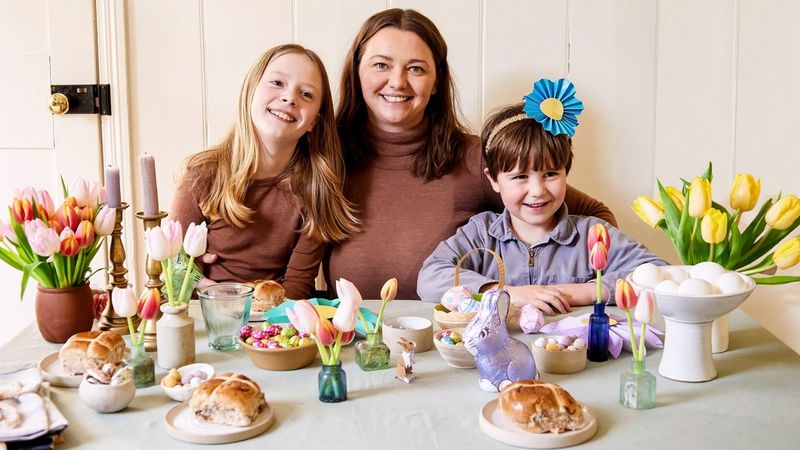 Crafter Hannah Bullivant sits between two children at a table decorated for spring with papercraft flowers, eggs and bunnies, vases of tulips and hot cross buns.