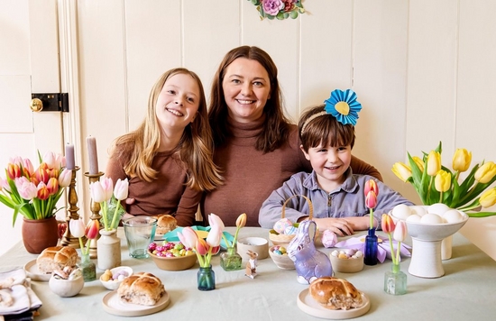 Crafter Hannah Bullivant sits between two children at a table decorated for spring with papercraft flowers, eggs and bunnies, vases of tulips and hot cross buns.