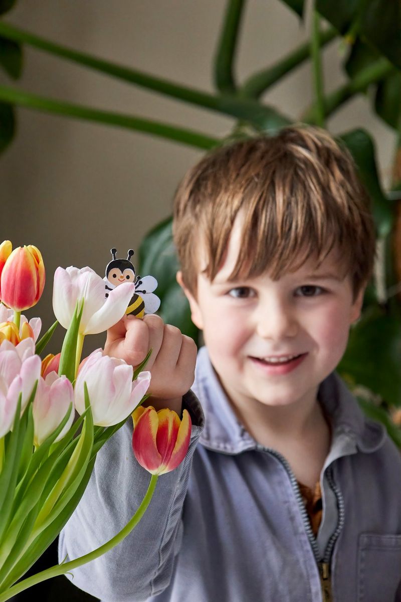 A young boy smiles as he holds a papercraft bee, made from a Creative Park template, alongside a vase of tulips.