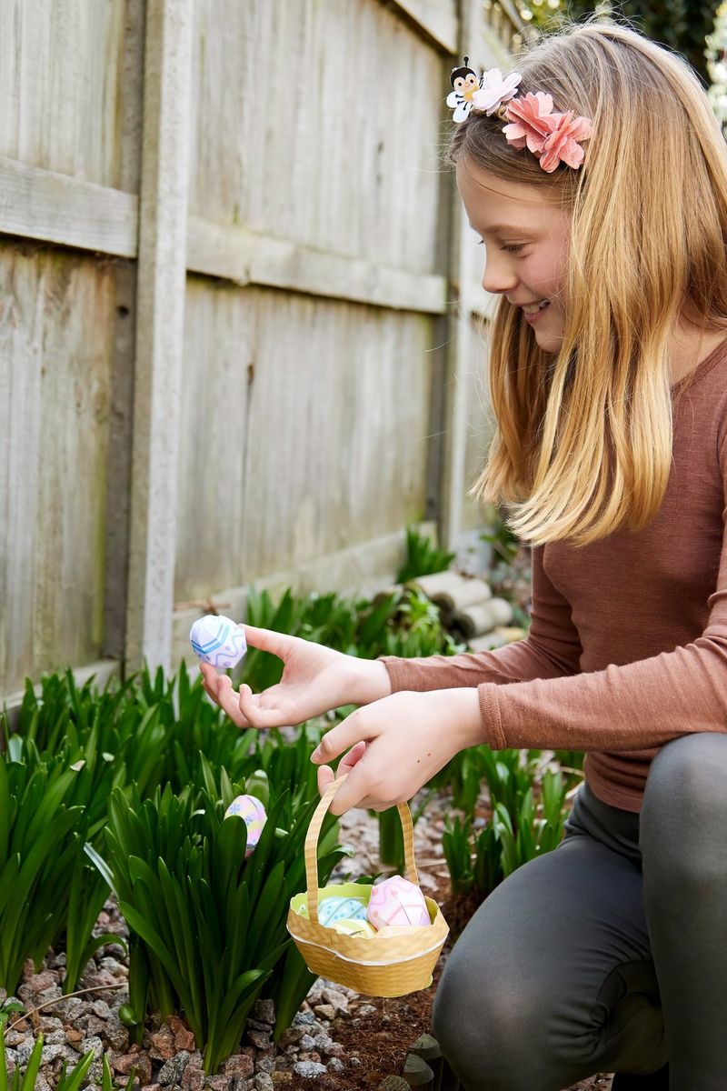A young girl kneels down alongside a flower border, holding a papercraft basket filled with papercraft eggs in one hand and a papercraft egg in the other.