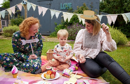 Two women holding papercraft photo booth props sit with a small child between them on a picnic blanket covered in colourful Creative Park papercrafts.