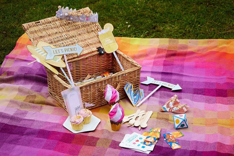 A picnic hamper filled with Creative Park papercrafts sits open on a colourful blanket. Surrounding it are several more papercrafts including paper ice creams, airplanes and a bingo game.