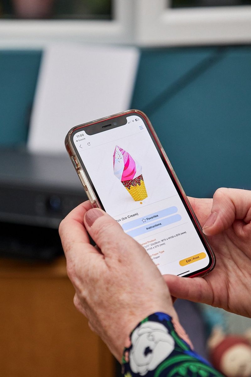 A pair of hands holding a smartphone displaying a papercraft ice cream in the Creative Park app.