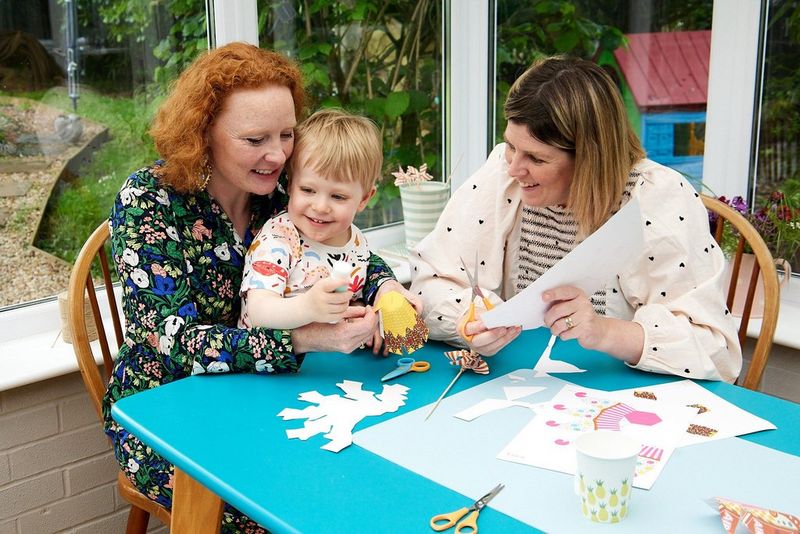 A young child sitting at a table between two women uses glue to stick a papercraft template of an ice cream together.