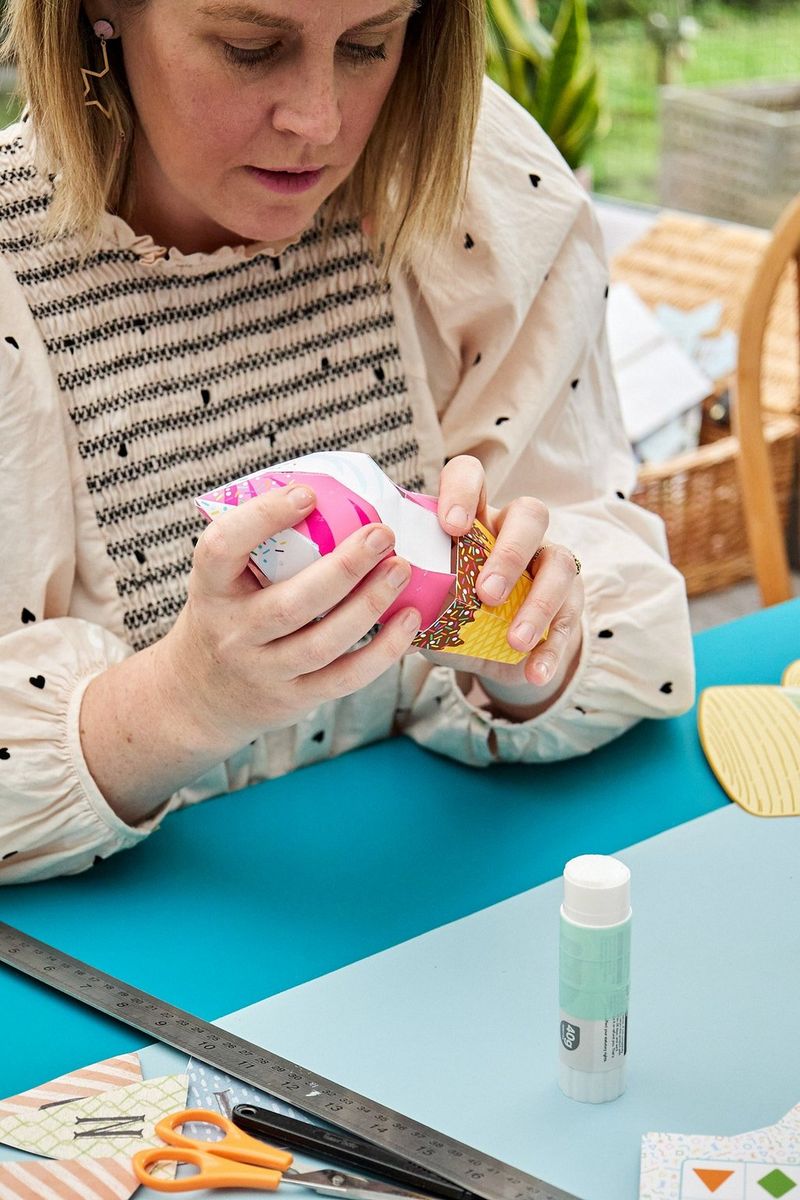 A woman sat at a small blue table in a conservatory carefully attaches two parts of a papercraft ice cream together.