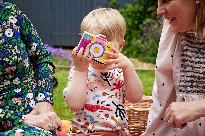 A young child sits in a garden between two women who are mostly out of frame holding a colourful papercraft camera up to his face.