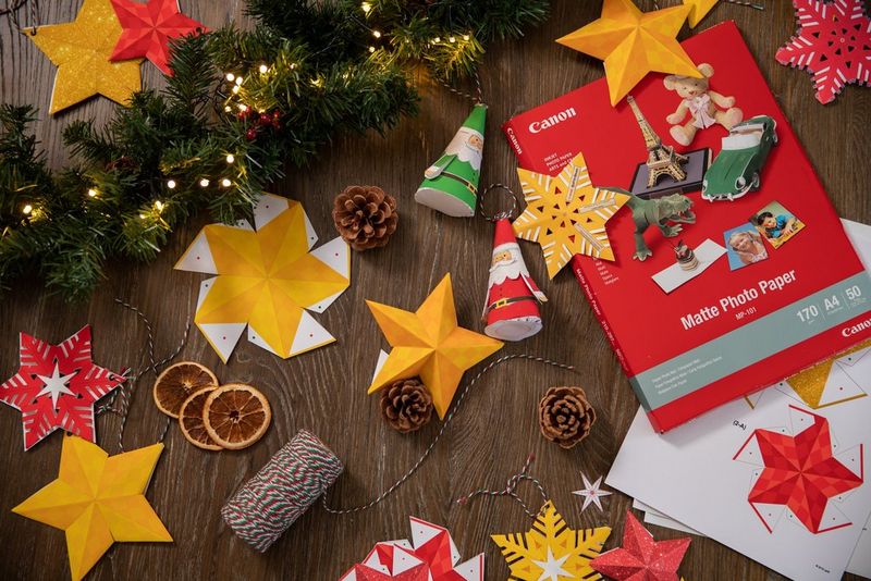 A flatlay image of a selection of papercraft Christmas decorations spread across a wooden table, alongside printed Creative Park templates, Canon printer paper, pine cones, twine and a festive branch.