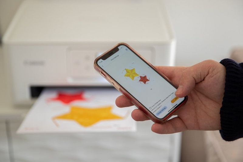 A hand holds up a smartphone showing a template for a papercraft Christmas decoration on the Creative Park app. In the background is a Canon PIXMA printer with a Creative Park template in the printer tray