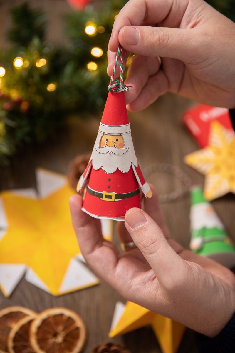 A pair of hands hold up a papercraft Santa Claus tree decoration attached to a loop of striped craft string.