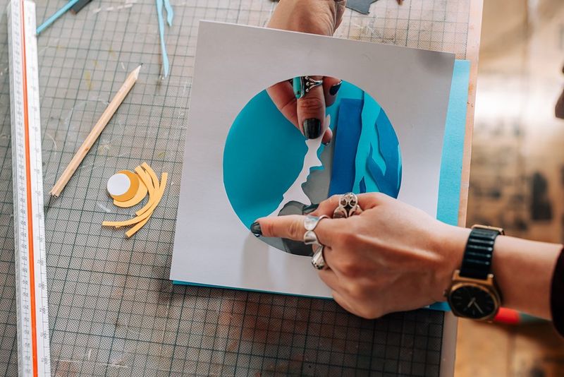 A person assembling a paper cutting piece, layering a cut-out of snow on top of some mountains. 