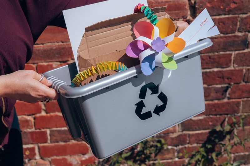 Hands hold up a grey recycling box, filled with pieces of cardboard and paper and discarded papercraft projects.