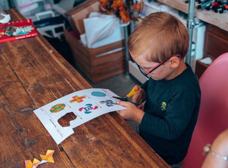 A young boy sits at a table carefully cutting out colourful shapes from a Creative Park template.