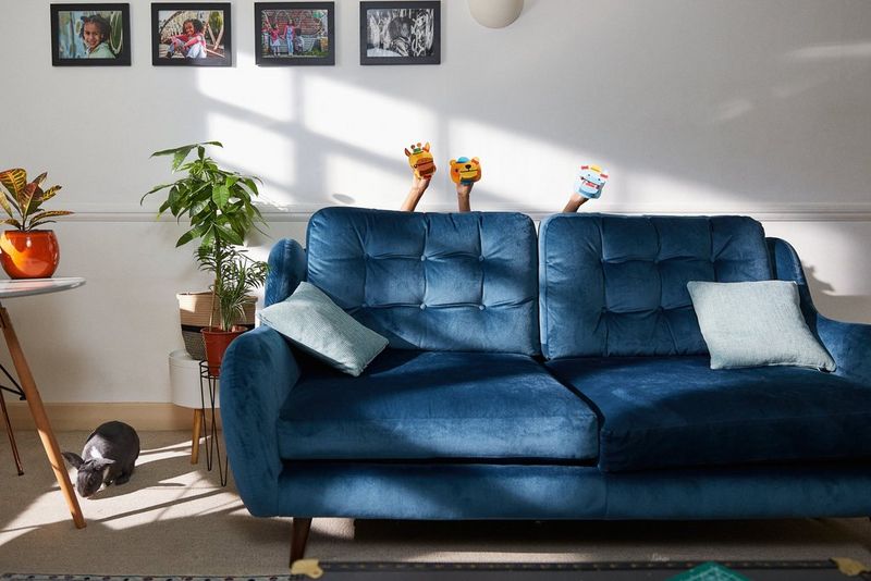 Three small hands hold up papercraft animal puppets from behind a large blue sofa in a family living room. To the left of the frame, a large rabbit can be seen.