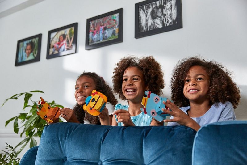 Three children behind a blue sofa, holding paper hand puppets and smiling.